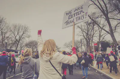 A woman holds up a placard as part of a protest in a public street. She is surrounded by other protestors. The placard reads 'we are better than this!'